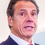 Photo of Andrew Cuomo, his face taut, likely in the middle of telling a bald-faced lie. He is wearing a blue jacket, white shirt, and red tie