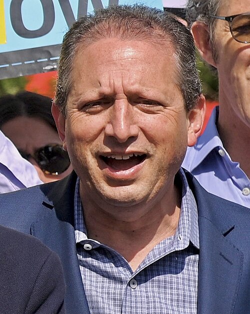 Photo of Brad Lander, mouth agape, in the midst of what looks like a rally. He is wearing a blue jacket and a blue and white checked shirt with no tie