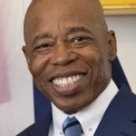 Photo of Eric Adams with a forced smile on his face. He's got on a blue tie, blue jacket, and a white shirt with a tie pin, which is really pretentious