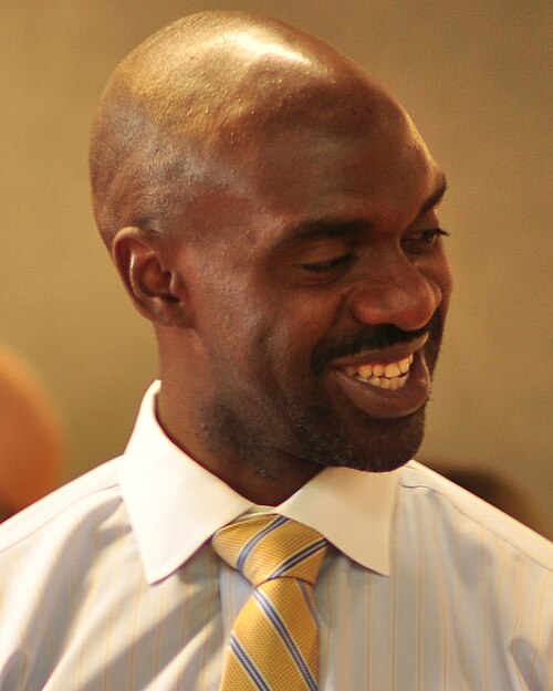 Photo of Michael Blake, smiling intensely as he looks down and to the right, wearing a white shirt and yellow and blue tie