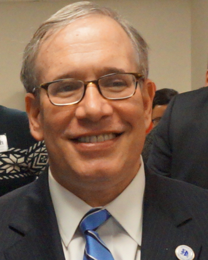 Photo of Scott Stringer looking straight-on and smiling while wearing a navy jacket, white shirt, and bright blue tie