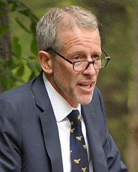Photo of Whitney Tilson looking over the top of his glasses frames while talking. He is wearing a navy jacket, white shirt, and navy tie with some design on it