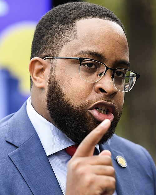 Photo of Zellnor Myrie in the middle of making a point. You can tell because his mouth is slightly parted and he is pointing into the air. He is wearing a blue jacket with a lapel pin, a light blue or white shirt, and a red tie