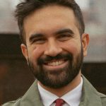 Photo of Zohran Mamdani, smiling up to his eyes. He appears to be wearing a green trench coat over a white shirt and red tie