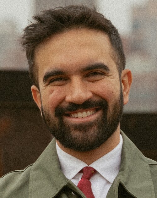 Photo of Zohran Mamdani, smiling up to his eyes. He appears to be wearing a green trench coat over a white shirt and red tie