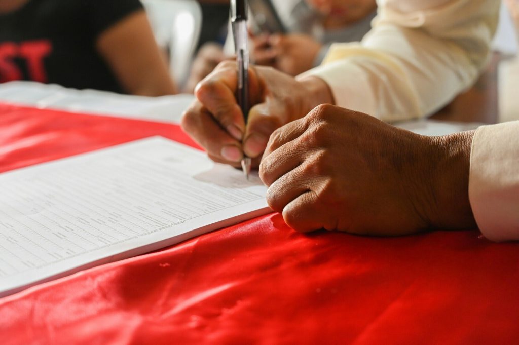 Image of a person filling out a form on a red-clothed table. Only their arms are visible, dressed in a cuffed shirt, holding a simple ballpoint pen. Other bodies can be seen, slightly blurry, in the background, one with a black t-shirt with red lettering