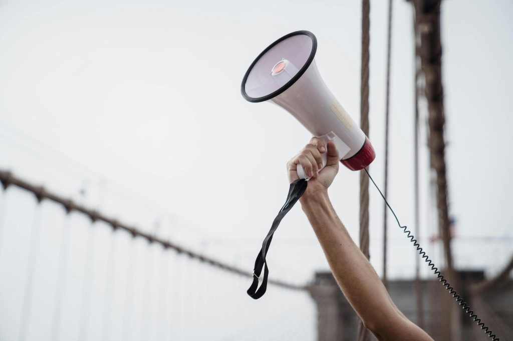 A photo of an arm holding a megaphone up in the air with the Brooklyn Bridge in the background
