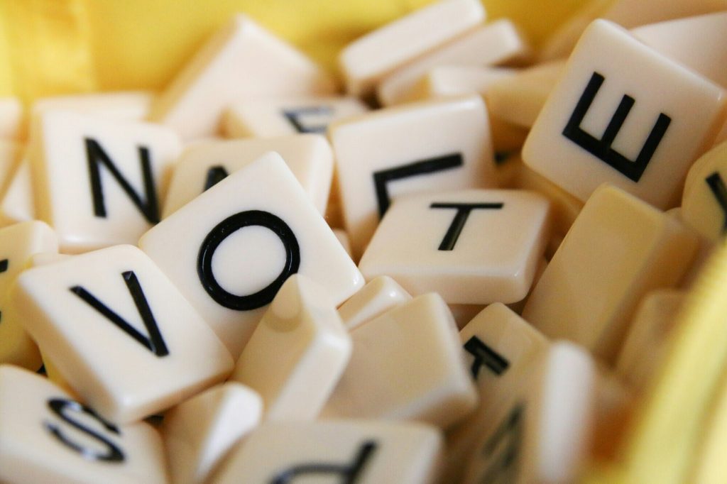 Image of a scrambled pile of Scrabble tiles. At the top of the pile are four clear tiles whose letters spell out the word "vote" (V-O-T-E)