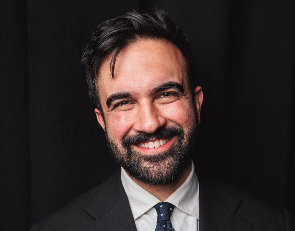 A photo of Zohran Mamdani, smiling casually, but with enough joy to wrinkle the edges of his eyes, wearing his typical simple suit and a blue tie with white dots