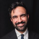 Image of Mayor Mamdani with a broad smile, clearly thinking of how amazing libraries are, in a dark suit and tie against a dark background