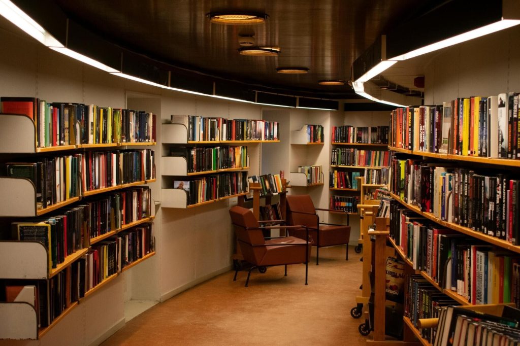 Photo of the interior of a library. There is a curving hallway lined with bookshelves and a couple of armchairs. The lighting is soft and gentle