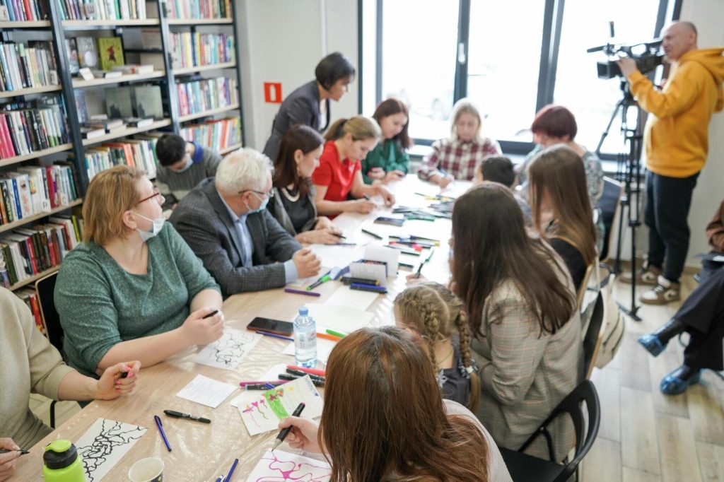 Photo of a group of people meeting around a table. Someone is videotaping the meeting and the back wall is covered by bookshelves. There is plenty of natural light streaming through a large window at the end of the table