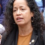 Photo of Alexa Avilés, a woman with long curly brown hair, a gray jacket and orange shirt. She looks calmly engaged in a policy she cares about sincerely