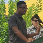 Photo of Chi Ossé speaking, with his hands, at a podium in front of some greenery and a yellow wall on a sunny day, with another person with short hair and sunglasses in the background