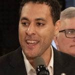 Photo of a light skinned man with short brown hair, speaking into a microphone with an energetic and gentle smile