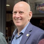 Photo of Harvey Epstein in a blue shirt and blue suit jacket, smiling in the sun