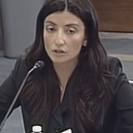 Photo of a white woman with long brown hair and dead eyes speaking into a microphone, probably about how we need more parking spaces and less affordable housing