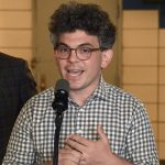 Photo of a white man with glasses and bushy hair, speaking into a microphone, seemingly animated by a real concern for his constituents. He's wearing a checkered shirt