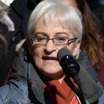Photo of a white woman in glasses with short, white hair, speaking before a microphone with clear thoughts and some sun in her eyes