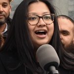 Image of an animated woman with long hair, eye glasses, and brown skin, speaking into a large microphone. She seems joyful, as though she's announcing some structural change to the city that will make her constituents' lives easier