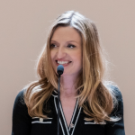 Photo of Virginia Maloney in a black and white top, speaking into a microphone, projecting gratitude