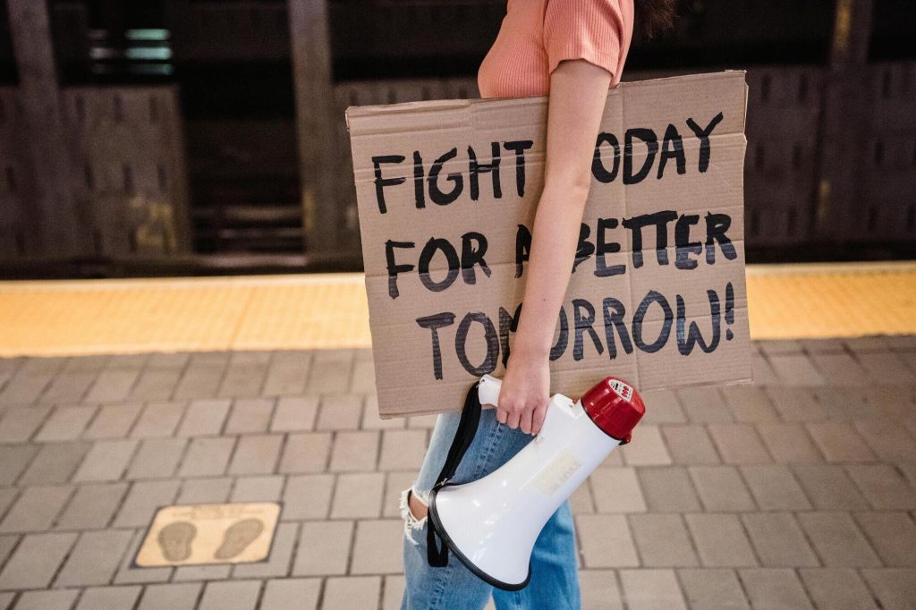 Image of someone standing on a subway platform holding a cardboard sign that reads "Fight today for a better tomorrow!" and a bullhort