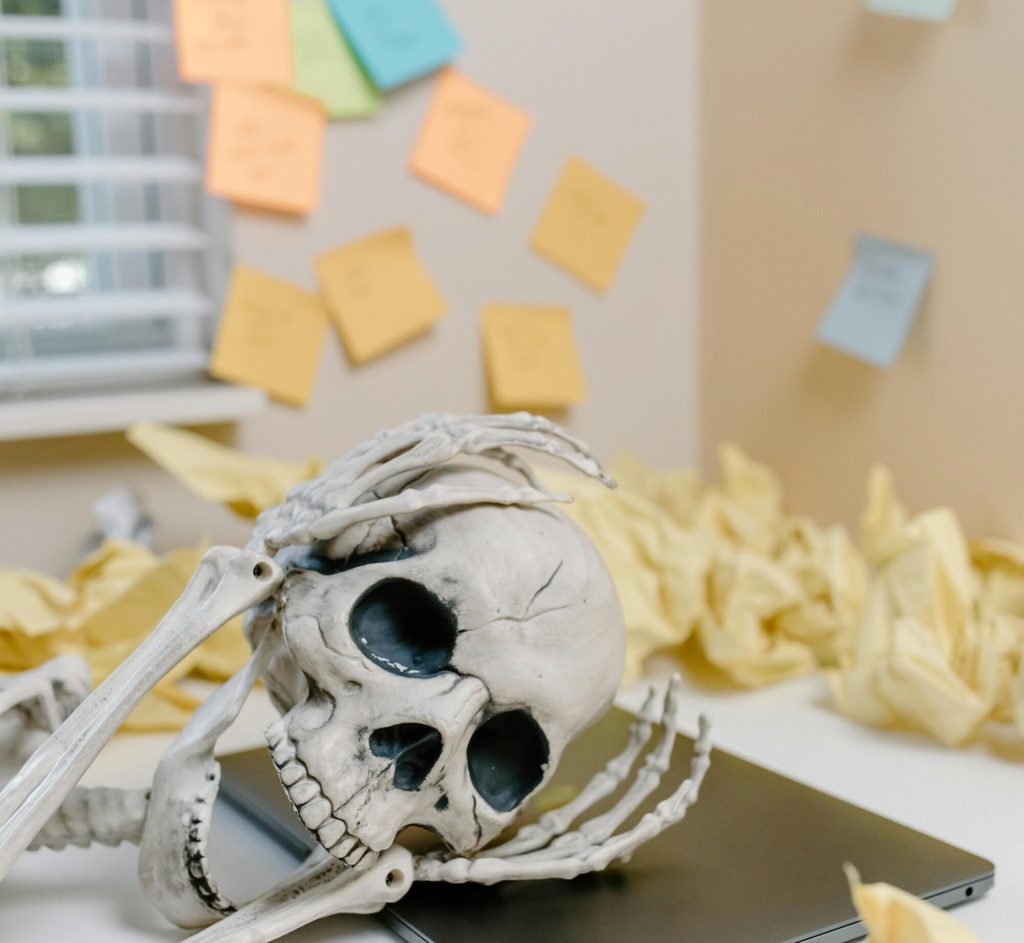 Image of a fake skeleton holding its head while leaning on a laptop at a desk in an office with tons of crumbled paper and sticky notes all over the walls in the background