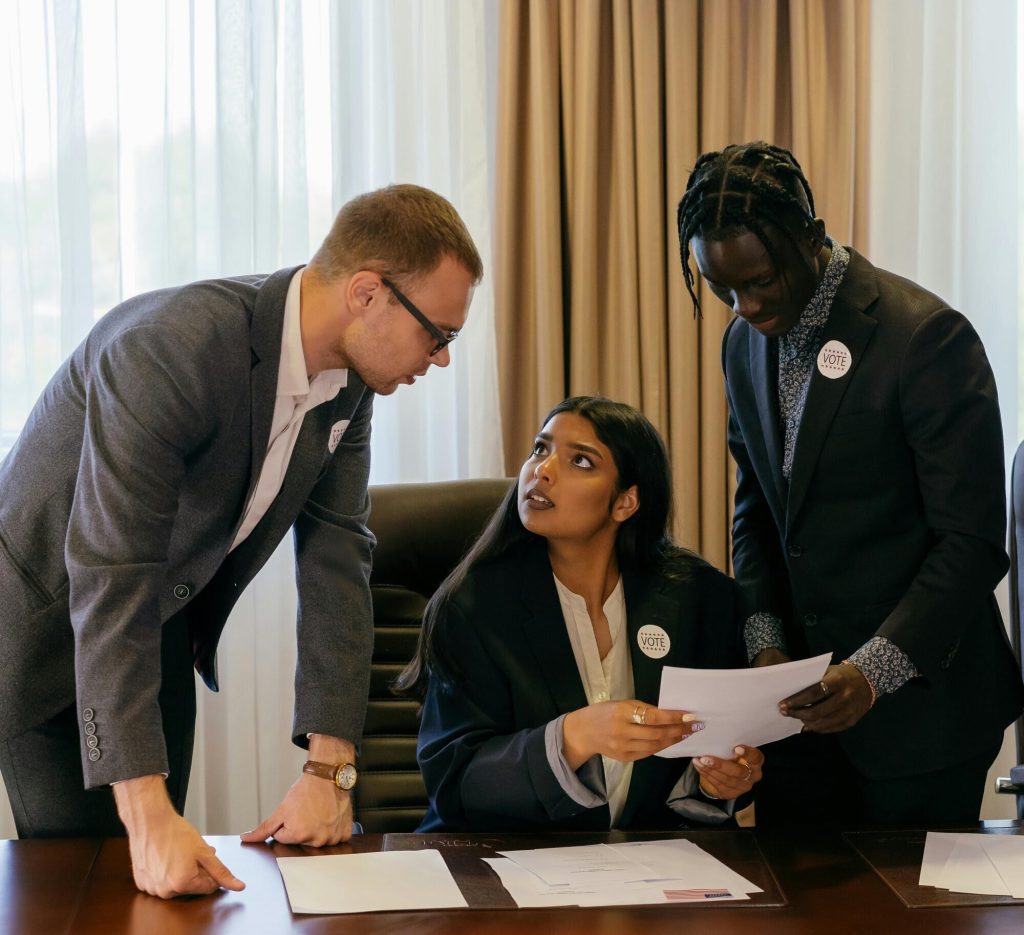 Photo of a woman sitting at a desk in a suit, apparently an elected official, with two men advising her. All three are wearing stickers that say "Vote"